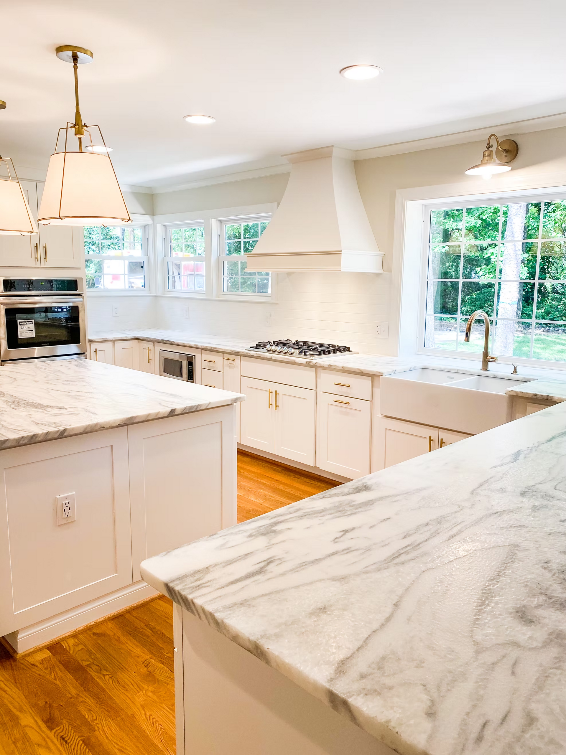 Bright white kitchen with marble countertops and wooden floors. Includes two islands, a farmhouse sink, and a range hood.