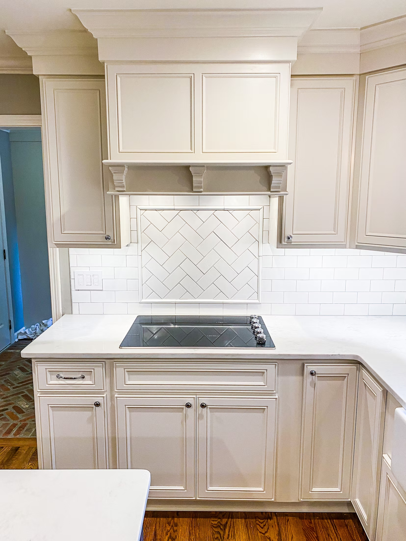 Cream-colored kitchen with a cooktop, herringbone backsplash, and upper cabinets. The counters and cabinets are white.