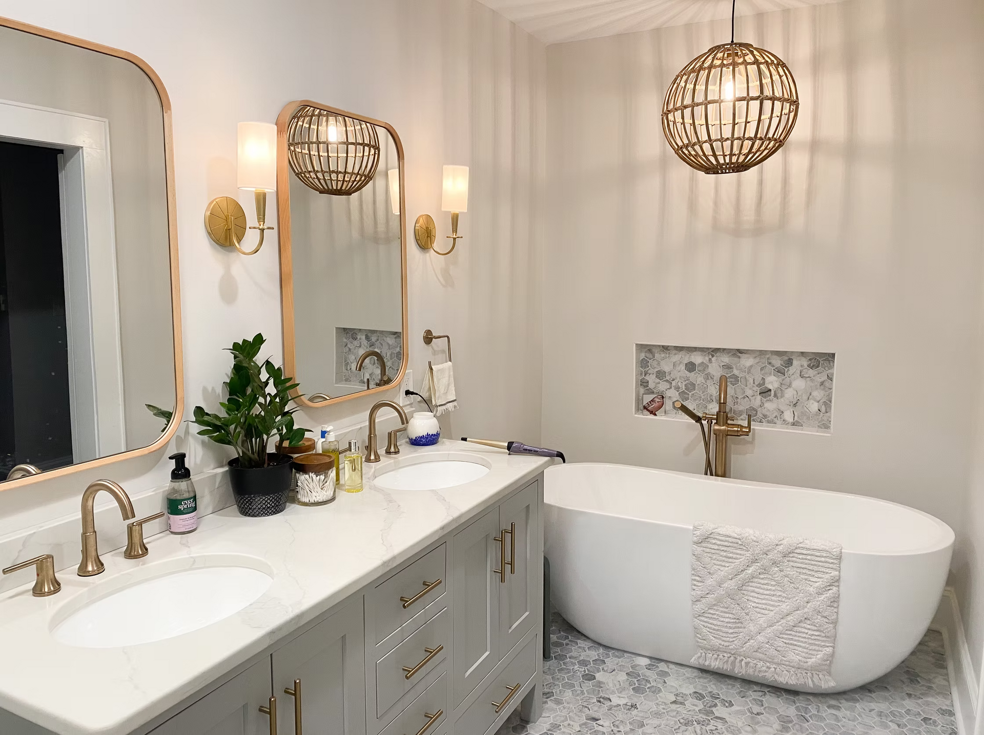 A modern bathroom with a freestanding tub, double vanity, and gold accents. The walls are neutral, and the lighting fixtures are decorative.