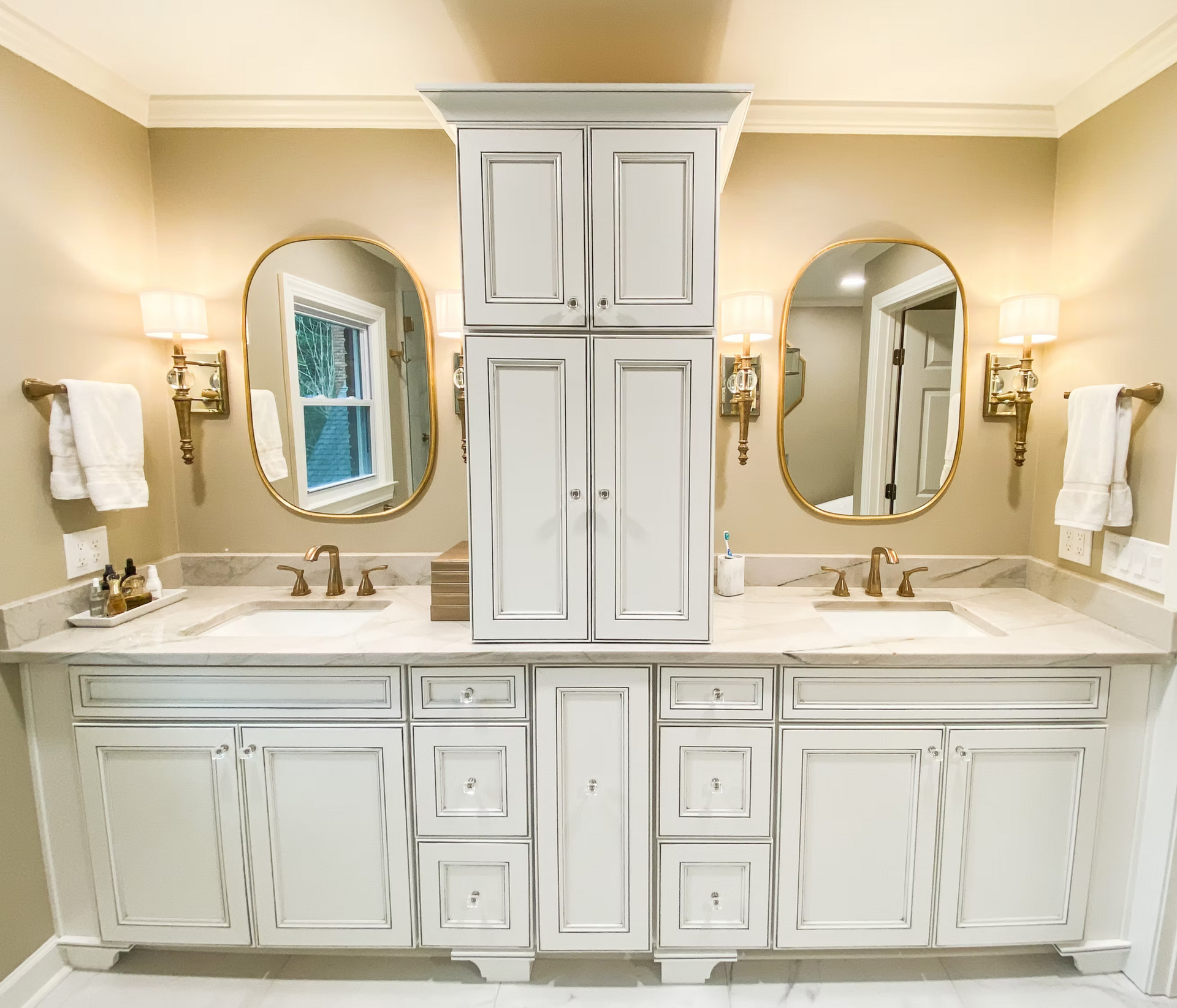 Bathroom with white cabinets, marble countertop, gold mirrors and sconces, and beige walls.  Double sinks and a tall cabinet are centered.