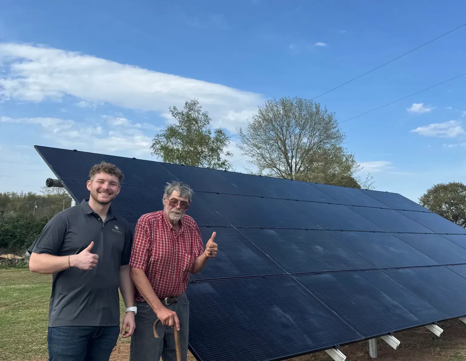 Two people smiling beside a large dark solar panel array outdoors under a blue sky