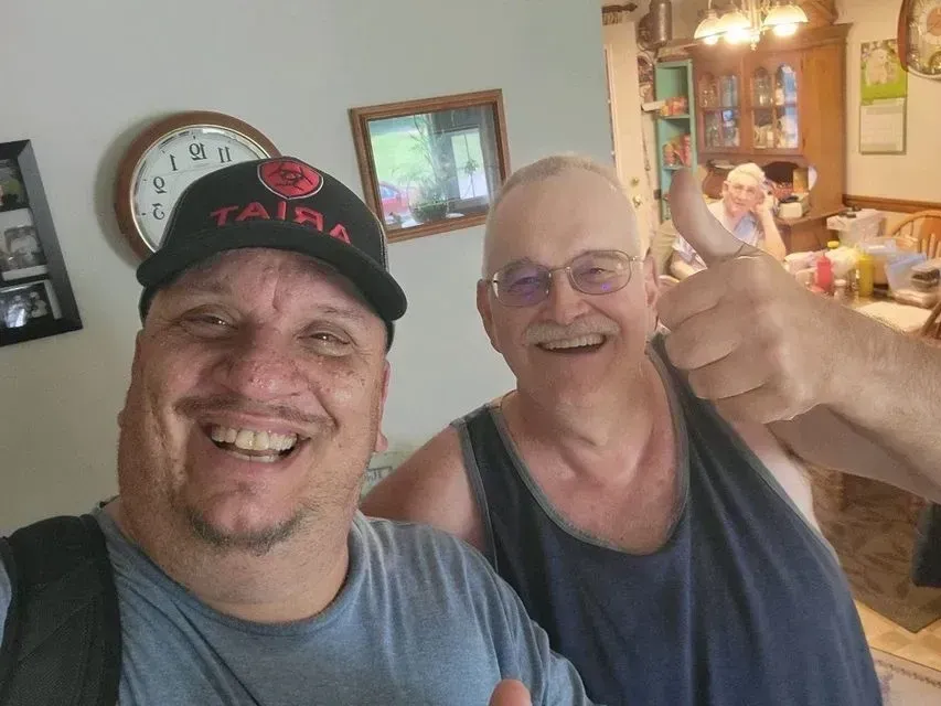 Two smiling men indoors, one taking a selfie and the other giving a thumbs-up.