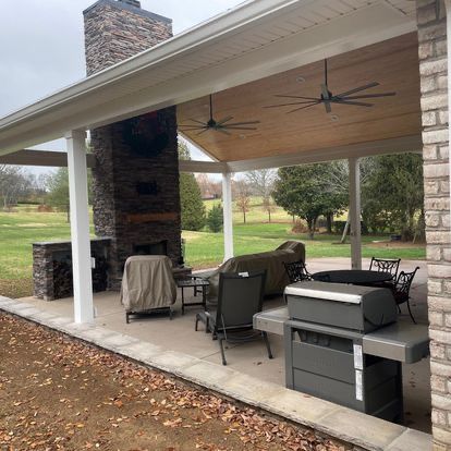 A covered patio with a grill , couch , chairs and a fireplace.