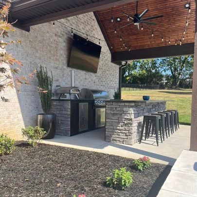 A patio with a grill, stools, a television and a ceiling fan.
