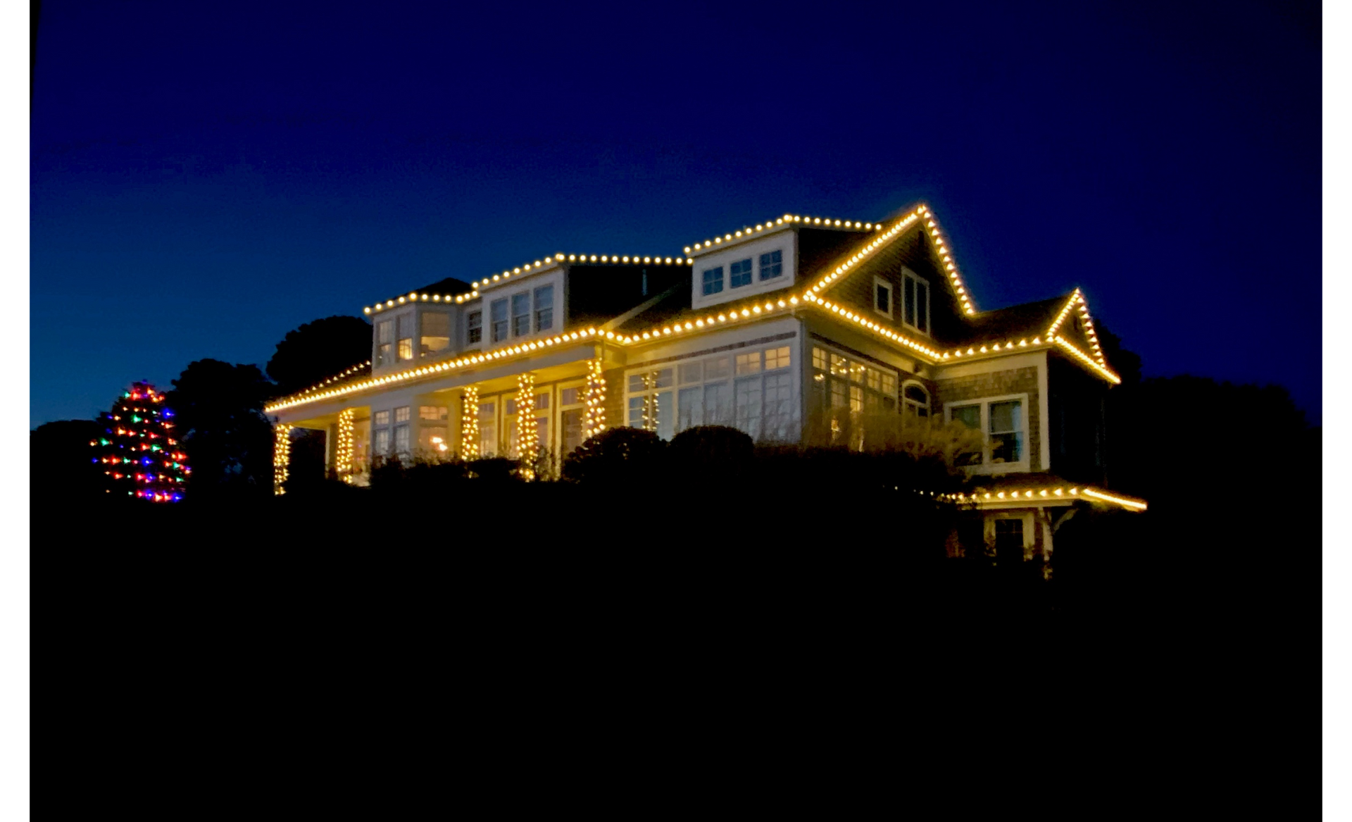 House at night, lit with yellow Christmas lights outlining the roof, windows, and porch.