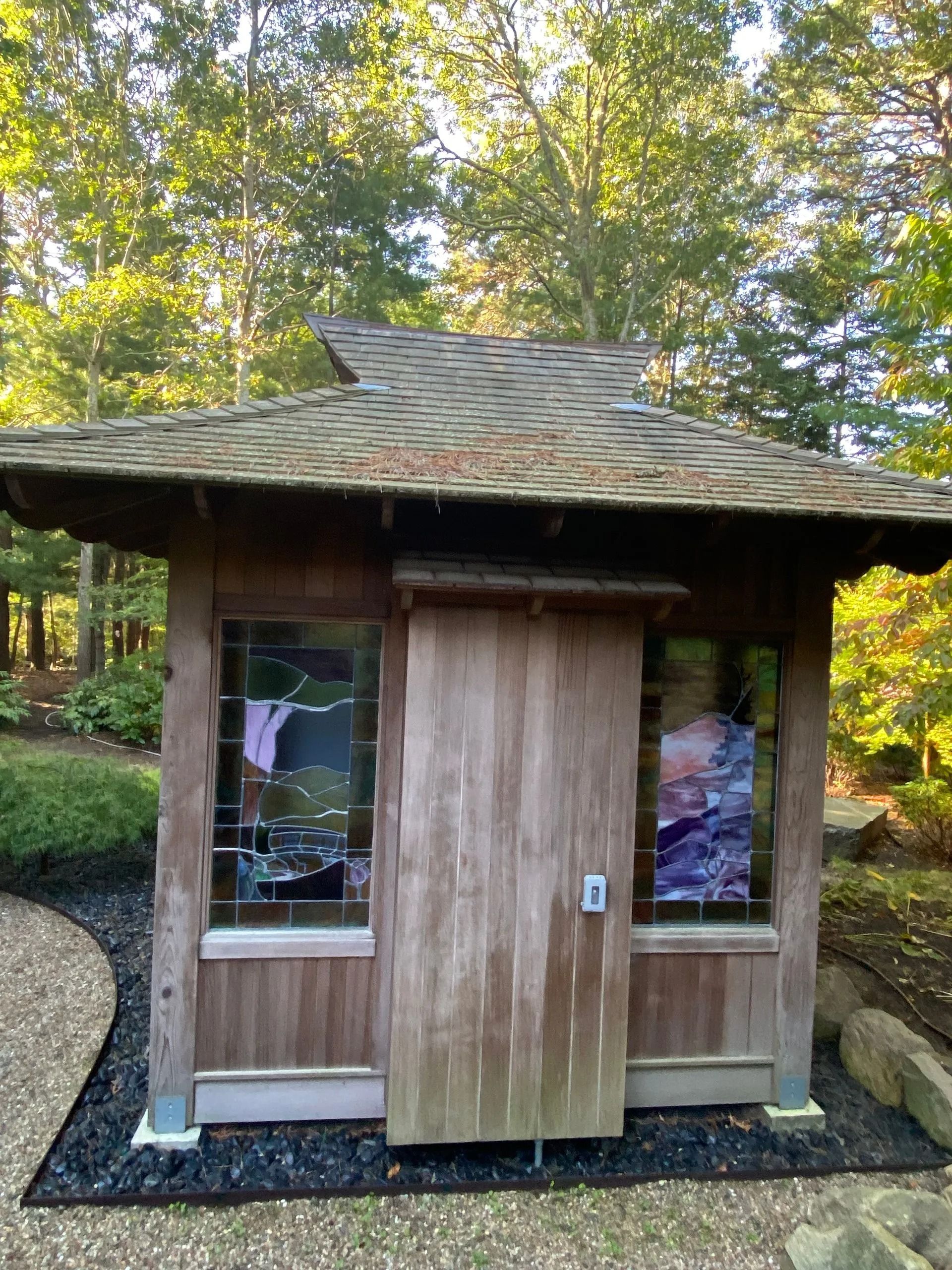Wooden Asian-style shed with stained glass windows, nestled in a garden with a curved path and trees.