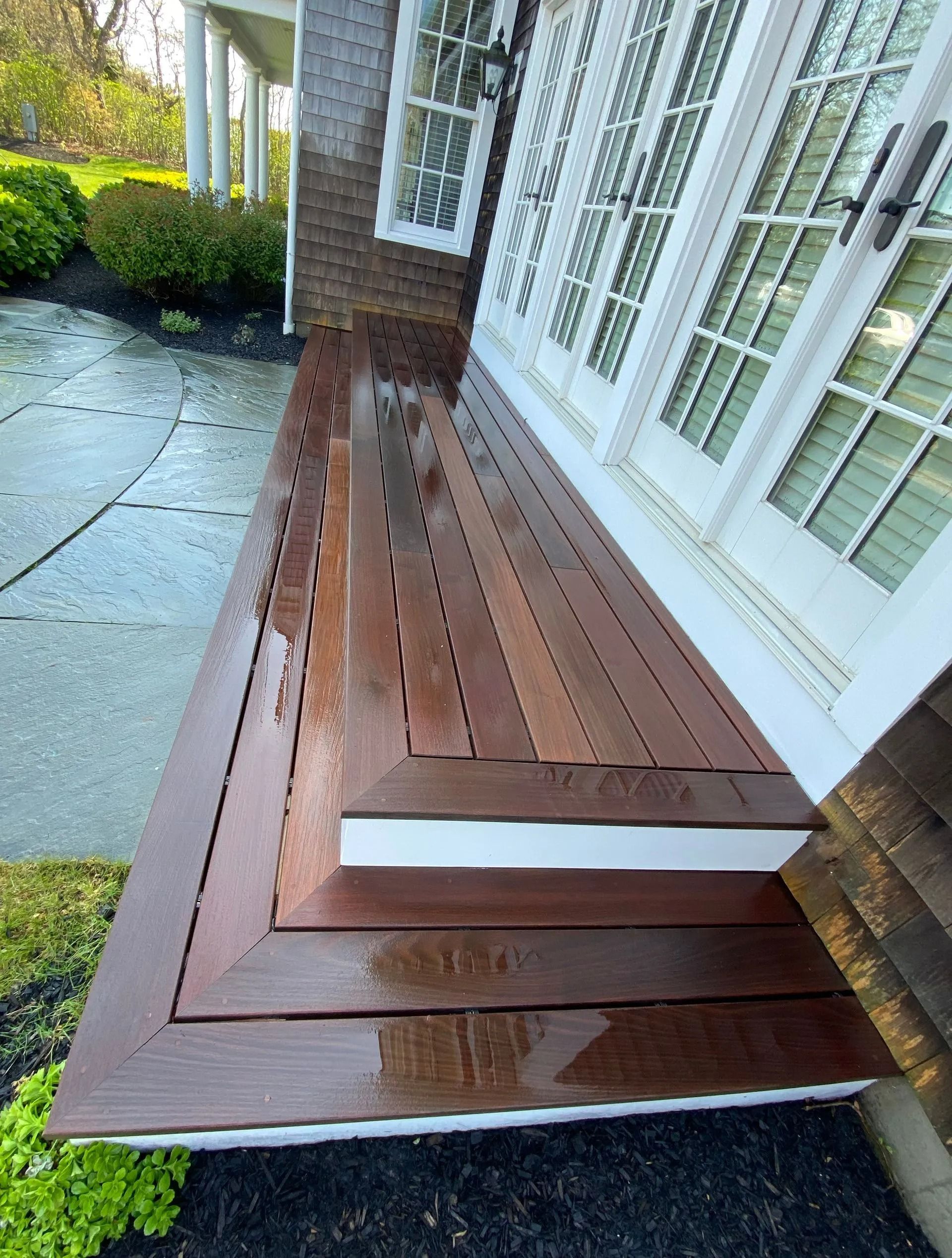 Wooden deck with steps, wet from rain, leading to white French doors. Brown wood, white trim.