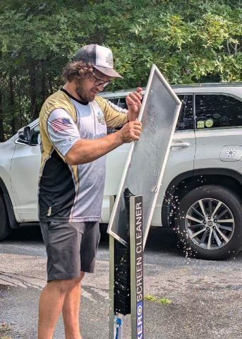 Man cleaning a screen outdoors; white SUV in background. He wears hat, grey shorts.