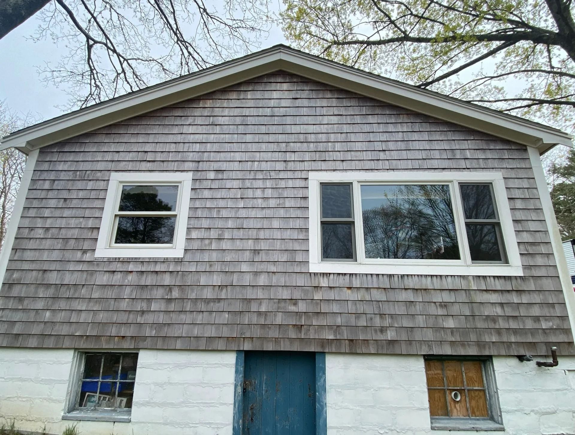 A two-story house with weathered wood shingles, white-painted brick foundation, and several windows.