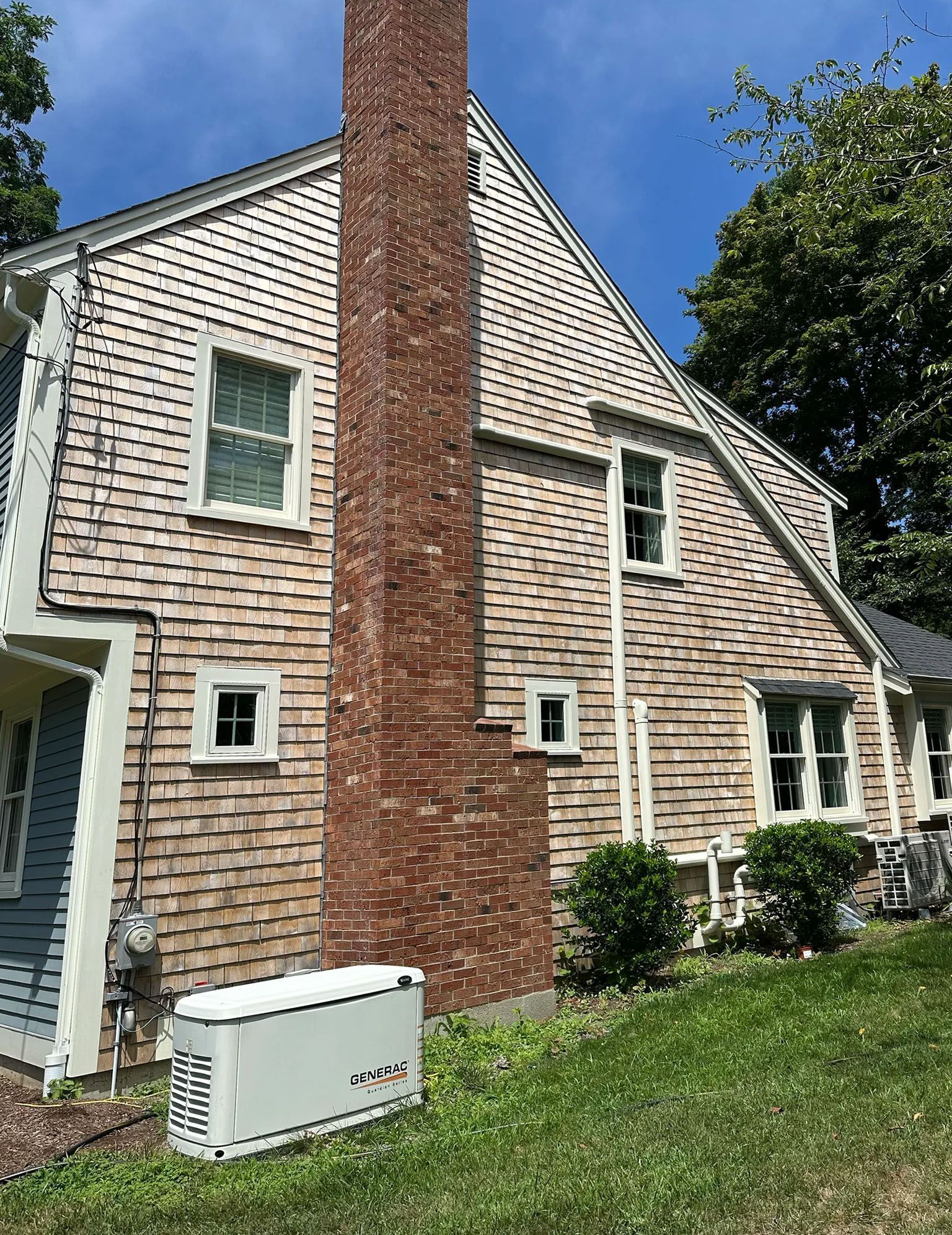 Side of a house with cedar shingles, a brick chimney, and white-framed windows. A generator sits below.