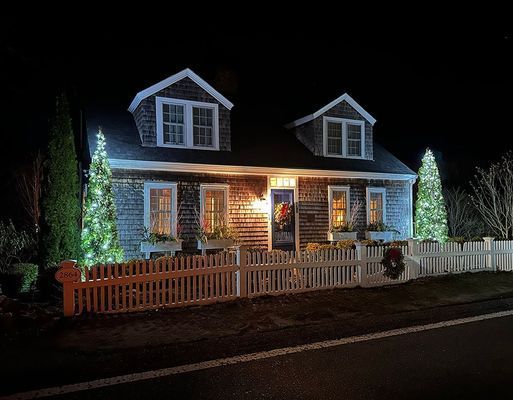 A quaint cottage at night with Christmas lights on the fence and house; two dormers, and a white picket fence.