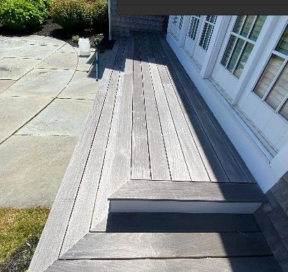 Wooden deck with steps leading to a house entrance. Gray wood, white trim, and sunlight.