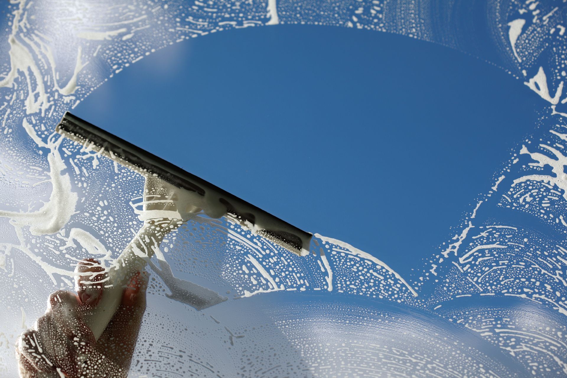 Hand squeegeeing soapy window, revealing clear blue sky.