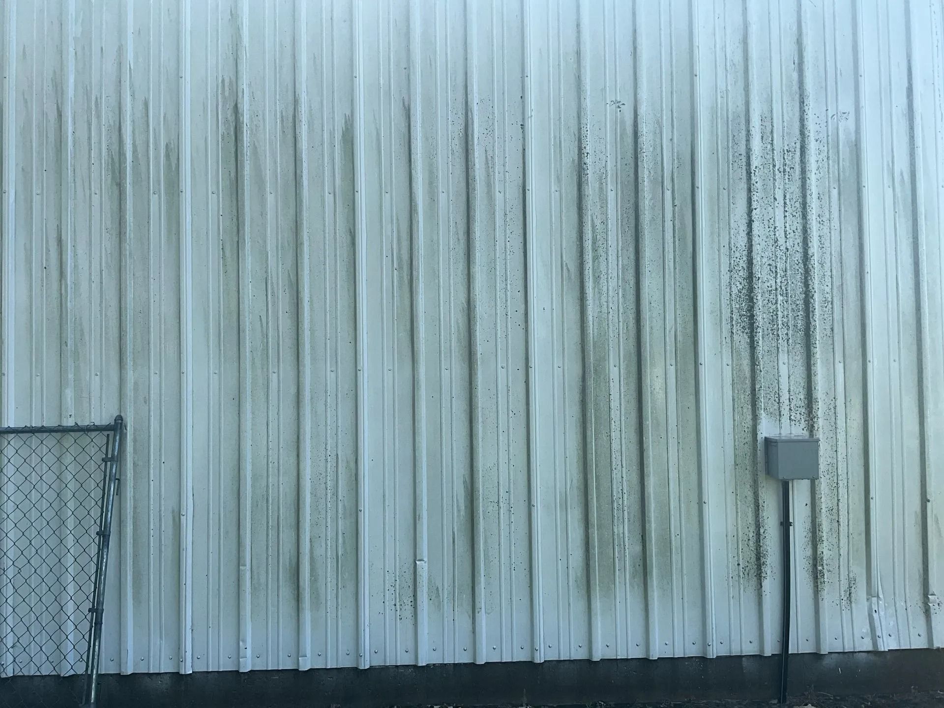 A white building with a dirty side and a chain link fence.