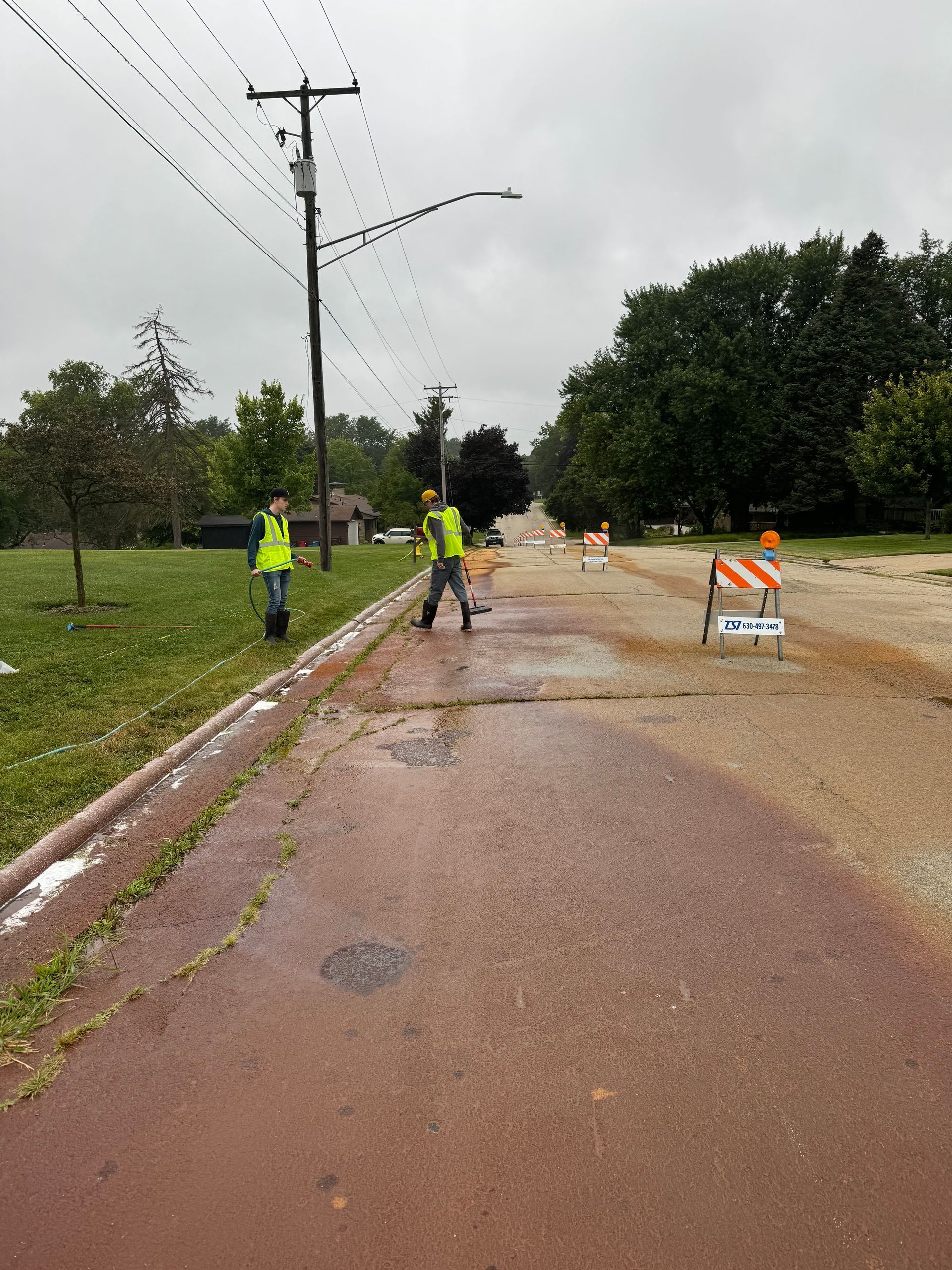 Two road workers in vests near a wet road with a barricade. Power lines and trees in background.