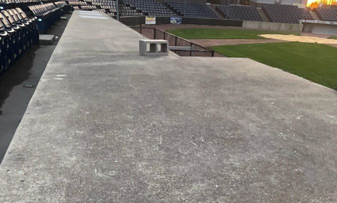 Concrete walkway alongside stadium seating, near the baseball field. A gray cinder block sits atop the walkway.