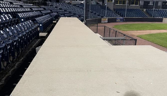 View from a concrete walkway, looking at blue stadium seats, baseball field, and fencing.