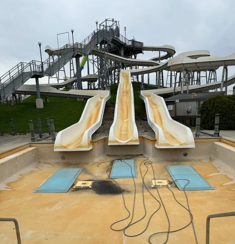 Three water slides with weathered white surfaces emptying into an empty, discolored pool. Metal stairs ascend to the slides.