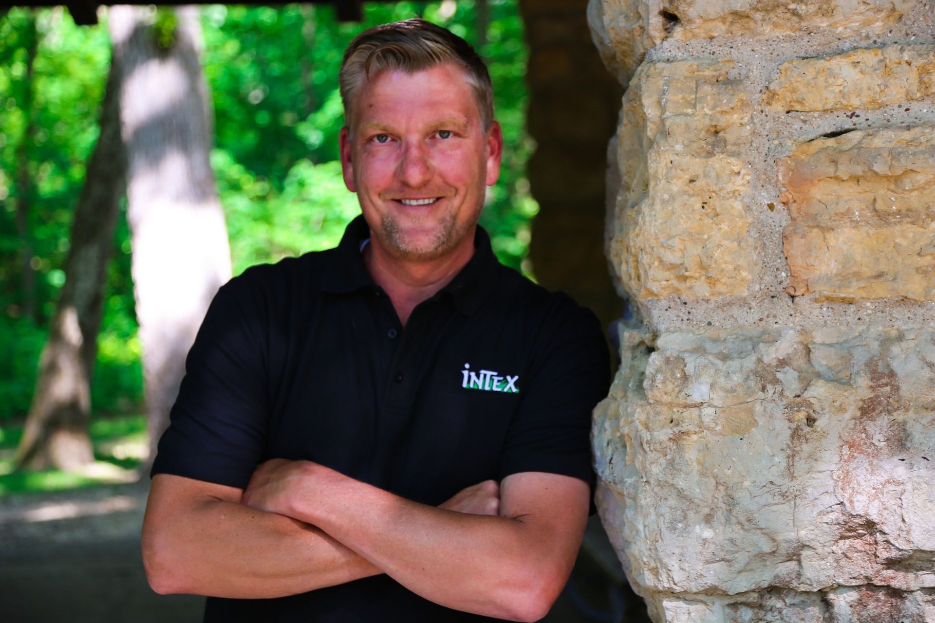Man in black shirt with arms crossed, smiling, leaning against a stone wall. Outdoors, natural light.