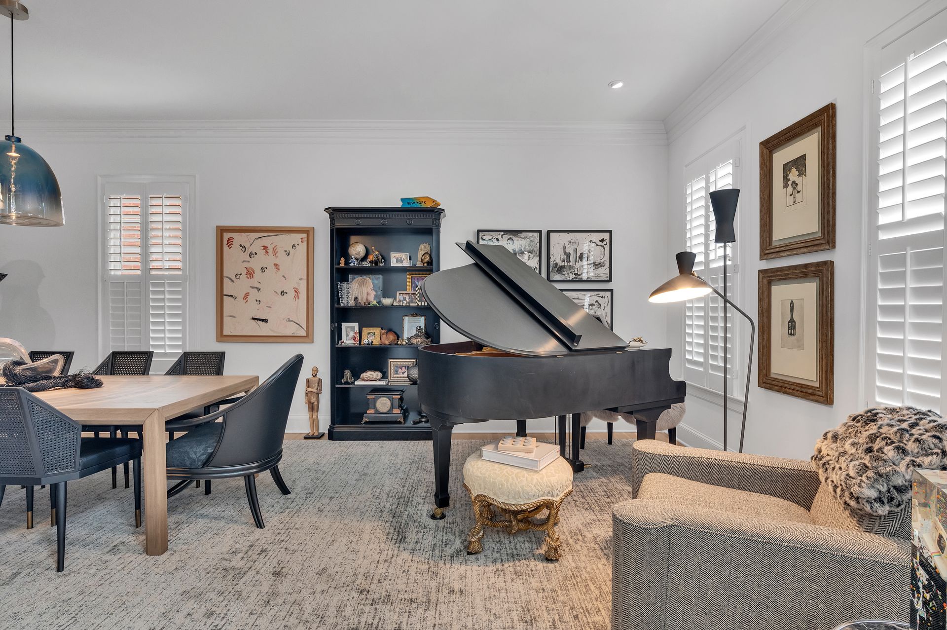 Living room with grand piano, dining table, and seating. White walls, wood shutters, and patterned rug.
