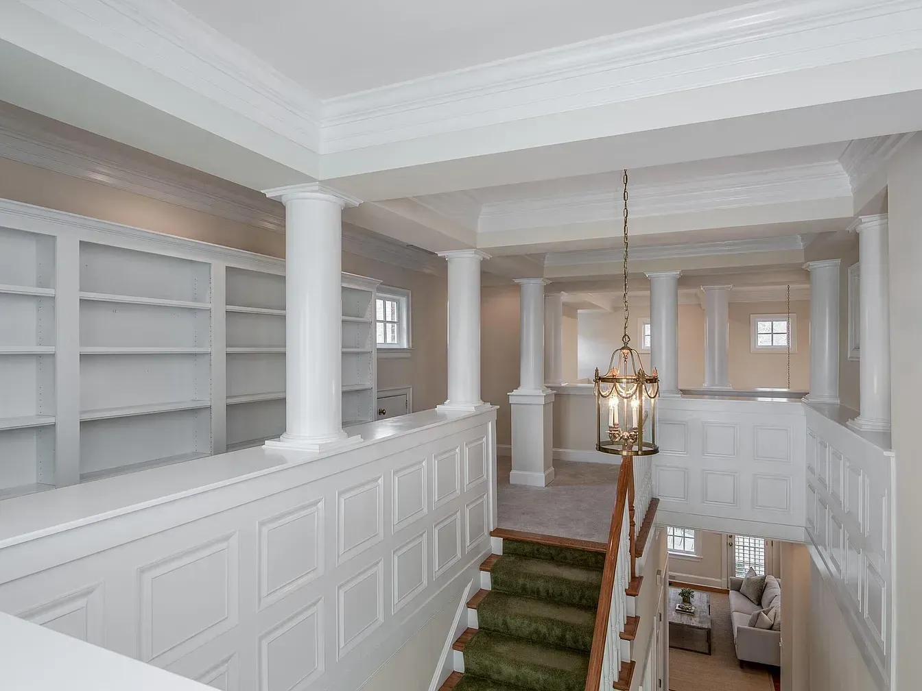 Interior view of a landing with white paneling, columns, staircase, and chandelier; empty bookshelves on the left.