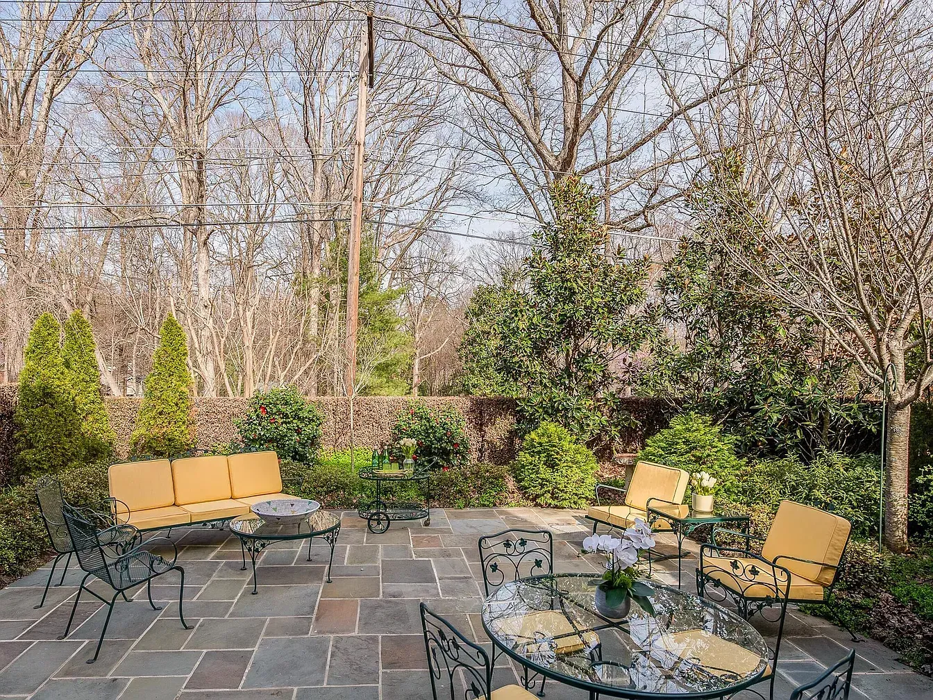 Patio with outdoor furniture, stone floor, and surrounding trees. Yellow cushions, glass tables, and metal chairs.