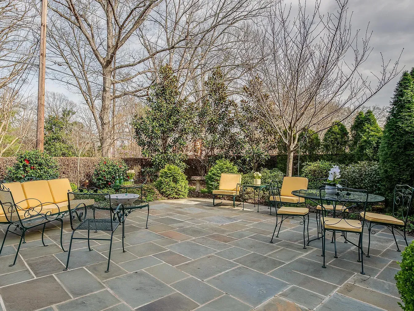 Patio with wrought iron furniture, surrounded by trees and bushes. Grey stone floor and yellow cushions.