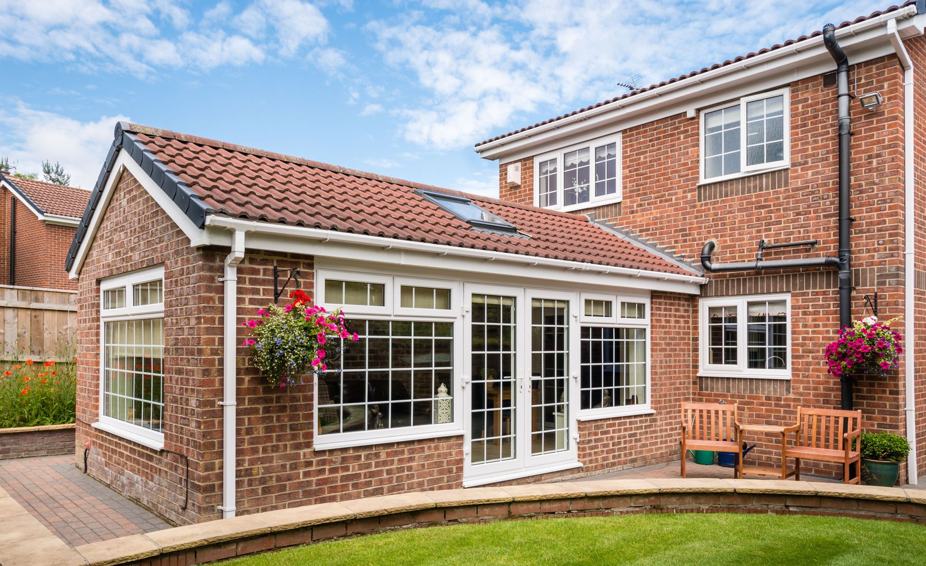 Brick home with an extension, featuring windows, doors, and a red tile roof. Patio with chairs and flowers.