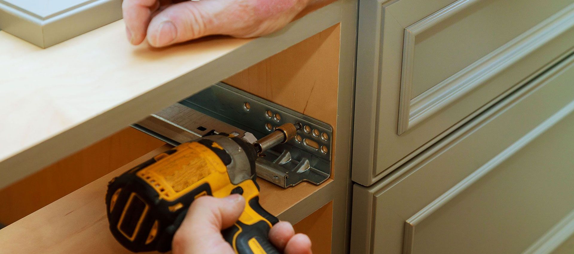 Hands using a yellow cordless drill to install a drawer slide in a kitchen cabinet.