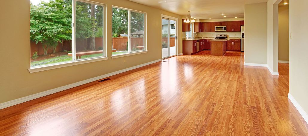 Empty sunlit living room with hardwood floors, large windows, and an open kitchen area.