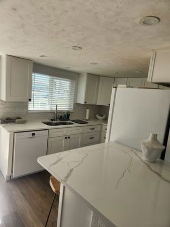 Bright white kitchen with marble island, cabinets, window blinds, and dark wood floor.