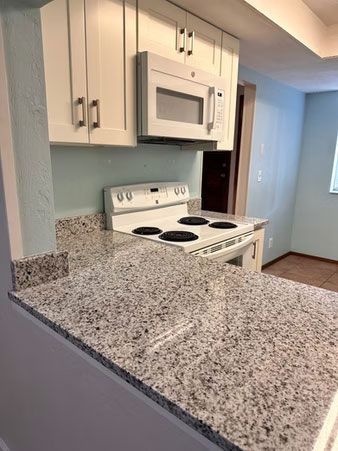 Small kitchen with white cabinets, speckled countertop, white stove, and microwave against light blue walls