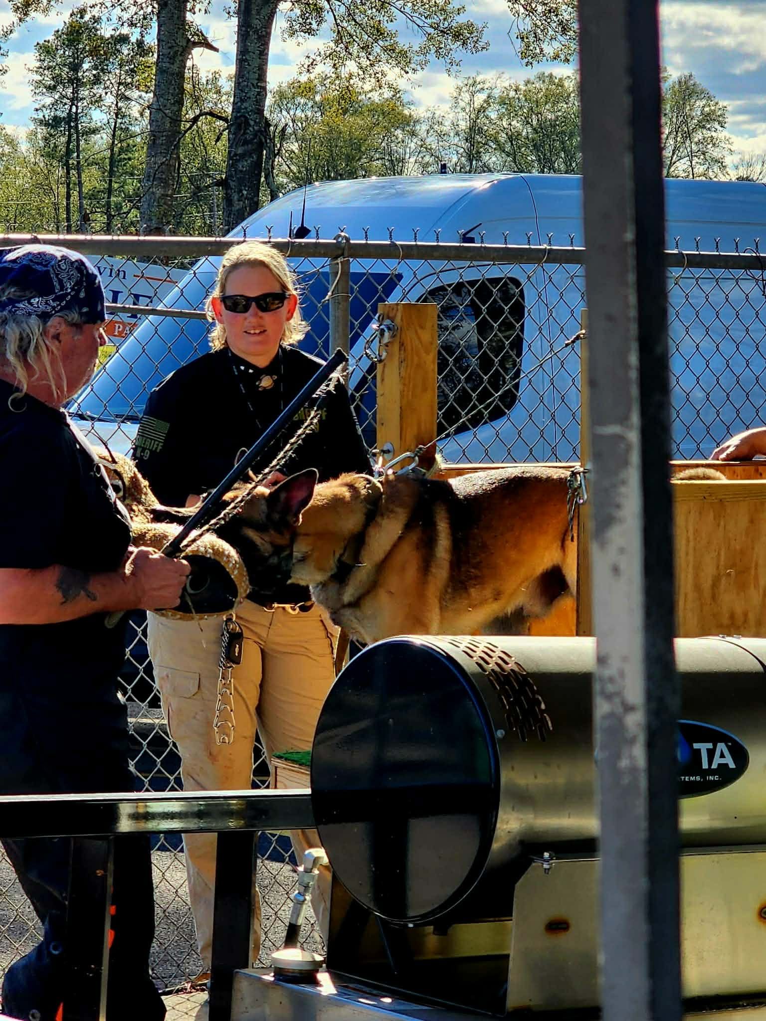 Two people with a dog, one holds a rifle, the other restrains the dog near a machine, outdoors by a fence.