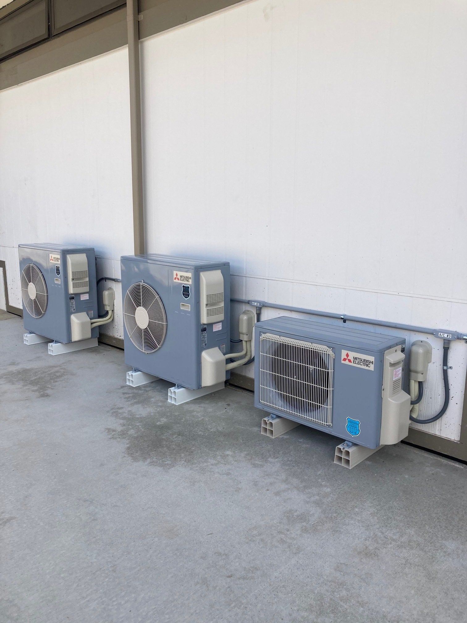 Three outdoor air conditioning units mounted on blocks against a white wall and gray floor.