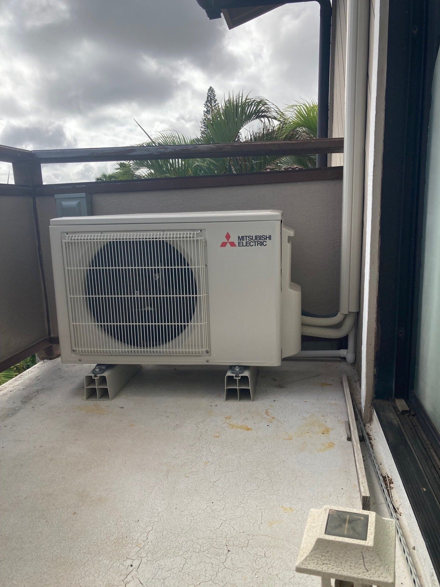 White Mitsubishi AC unit on concrete blocks, balcony setting. Cloudy sky.
