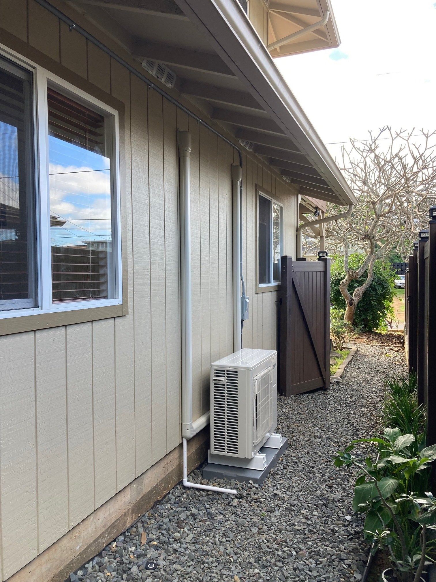Exterior of a beige building with windows, a brown door, and an air conditioning unit. Gravel path.