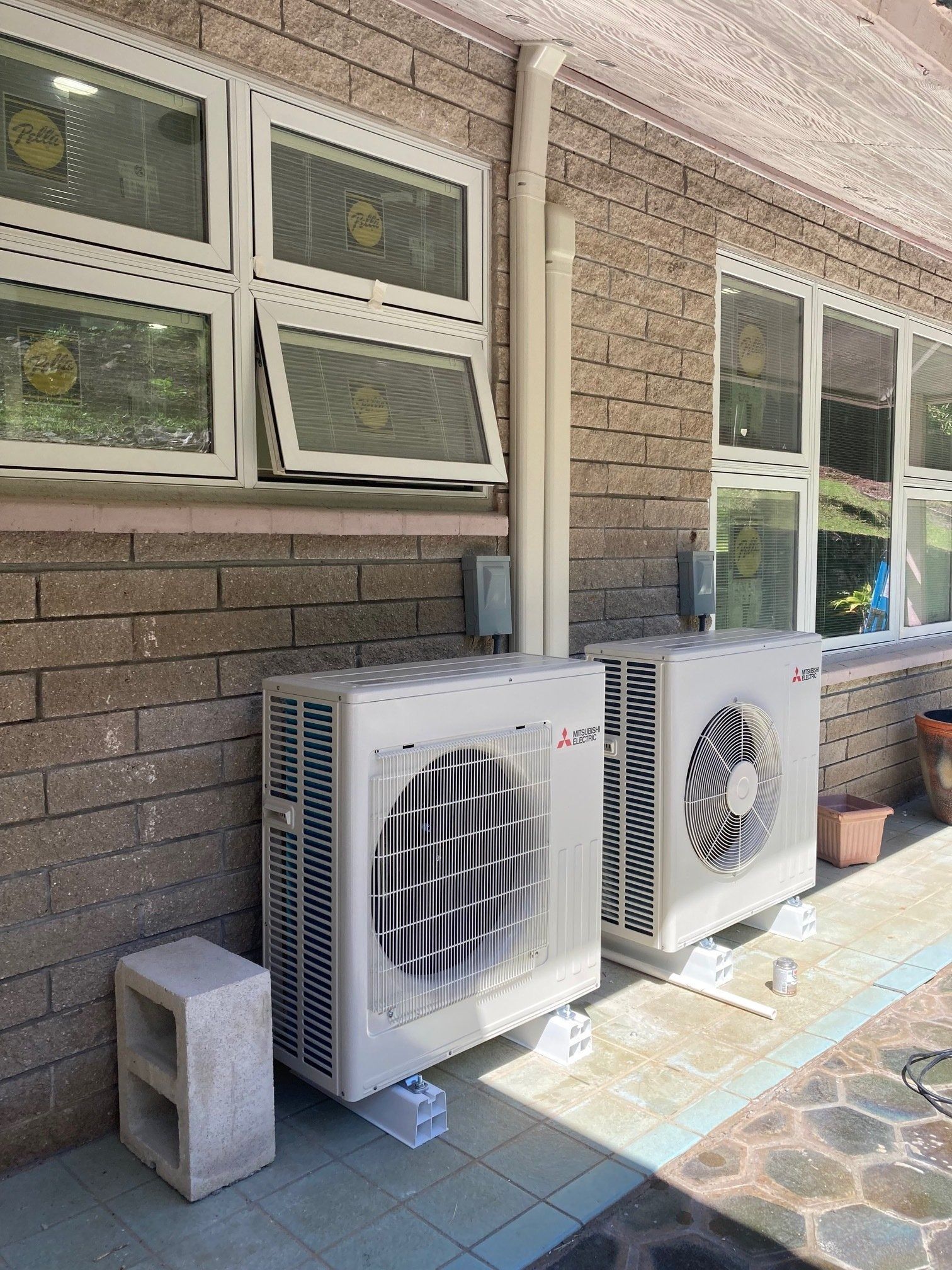 Two white air conditioning units outside, near windows on a brick wall. A concrete block is nearby.