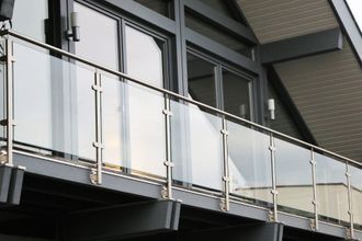 Glass balcony railing with metal posts, in front of windows of a building.