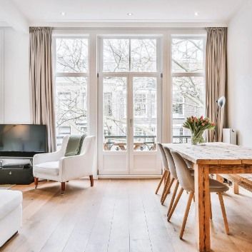 Living room with large windows, white armchair, dining table, hardwood floors, and beige curtains.