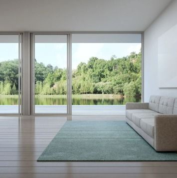 Modern living room overlooking lake and trees. Light beige couch on blue rug, large windows.