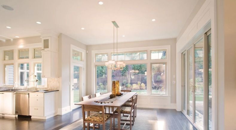 Bright dining room with wooden table, chairs, and large windows overlooking a yard.
