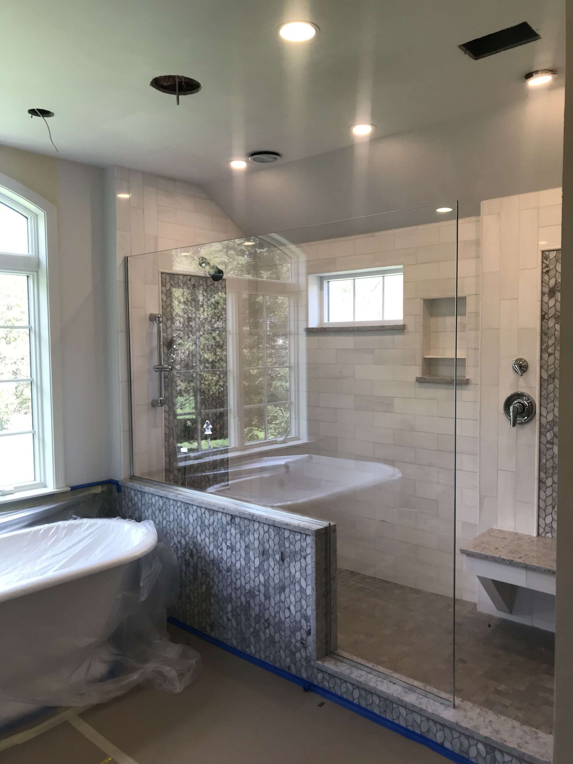 Spacious, light-filled bathroom under construction with a glass shower, tub, and window. Gray and white tiles.