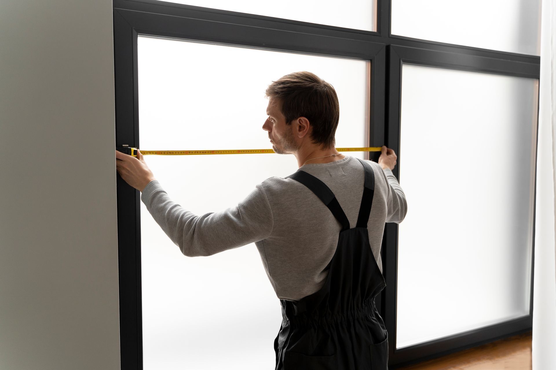 Man in overalls measuring a window with a tape measure in a room.