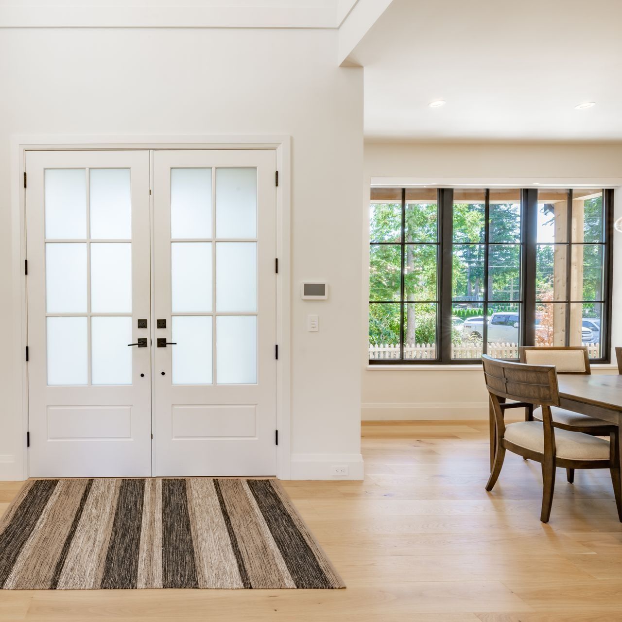 White interior with double doors and a striped rug, leading to a dining area with large windows.