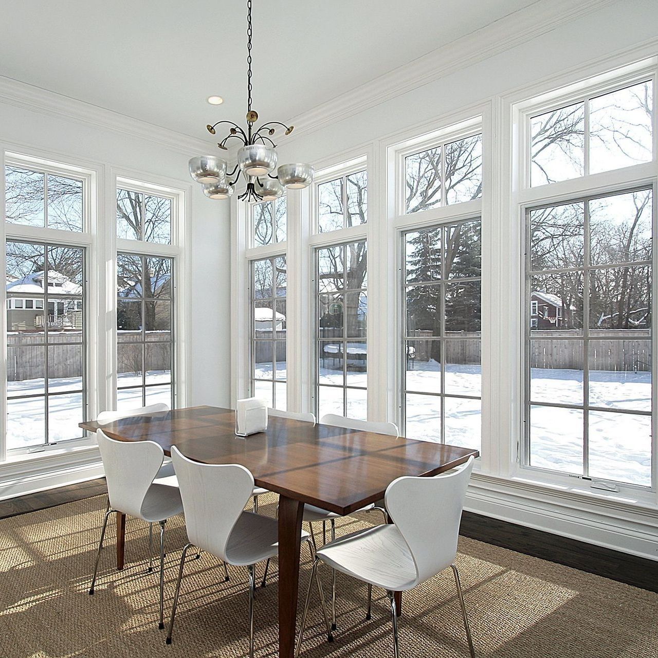 A bright dining room with a wood table and white chairs by large windows looking out at a snowy yard.