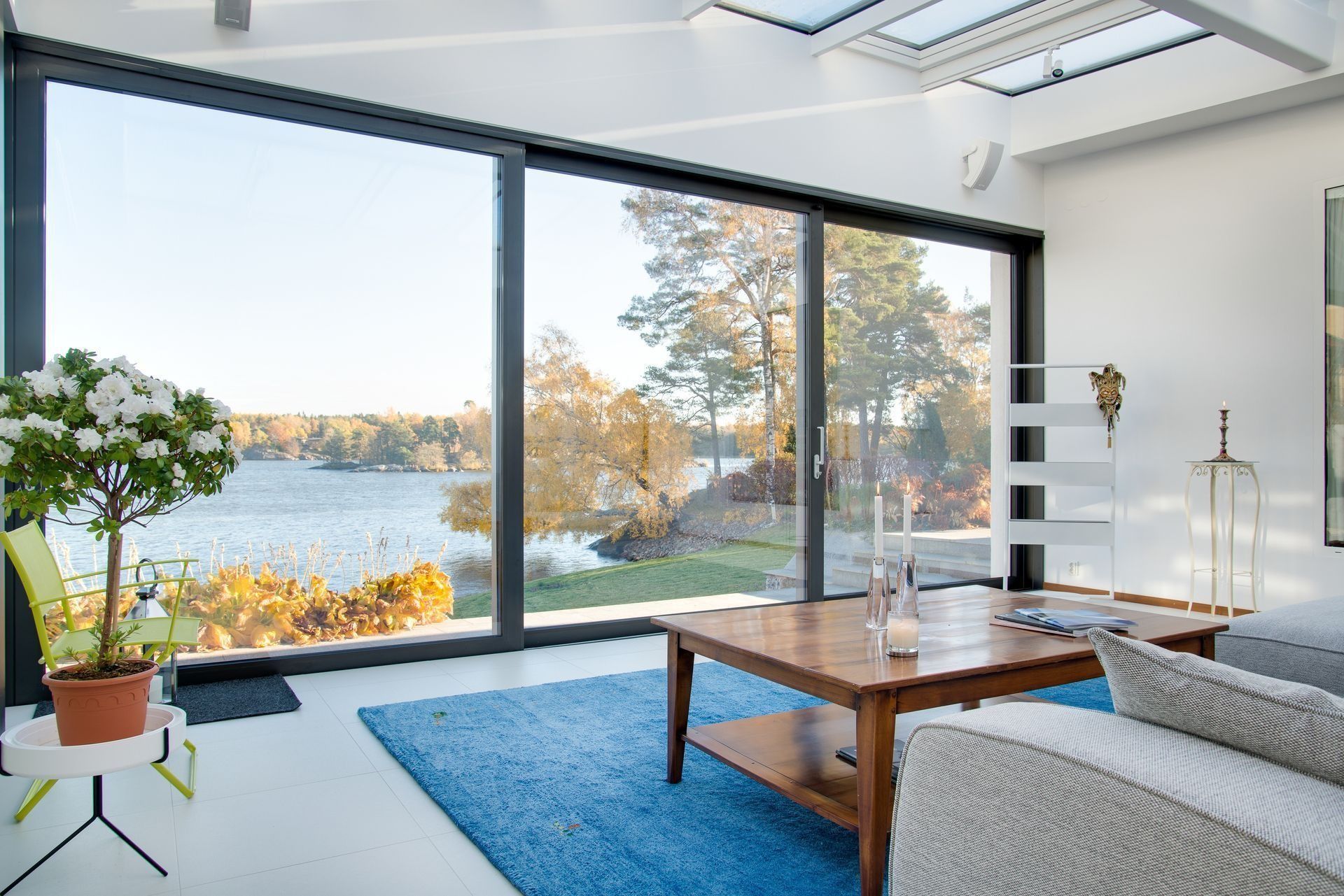 Living room with large windows overlooking a lake, featuring blue rug, wooden coffee table, and potted plant.