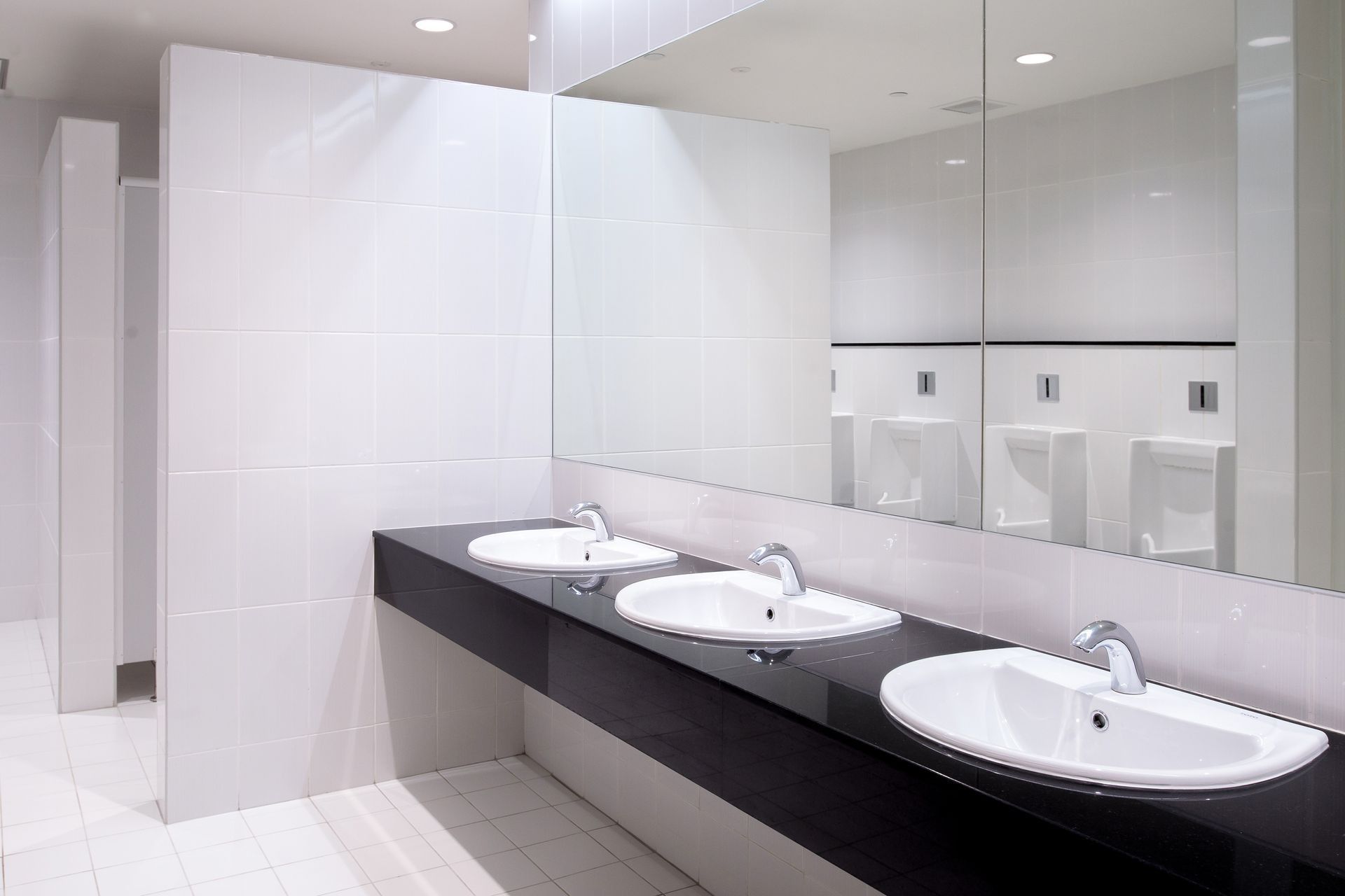 Public restroom with three sinks, large mirror, and urinals. White and black tones.