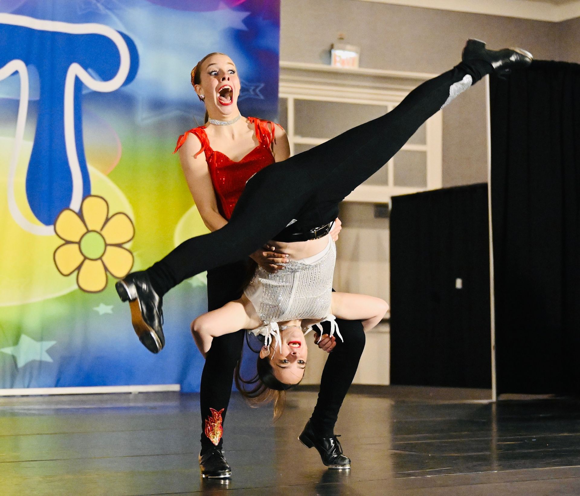 Two female tap dancers performing a lift on stage. One is upside down, while the other is supporting her.