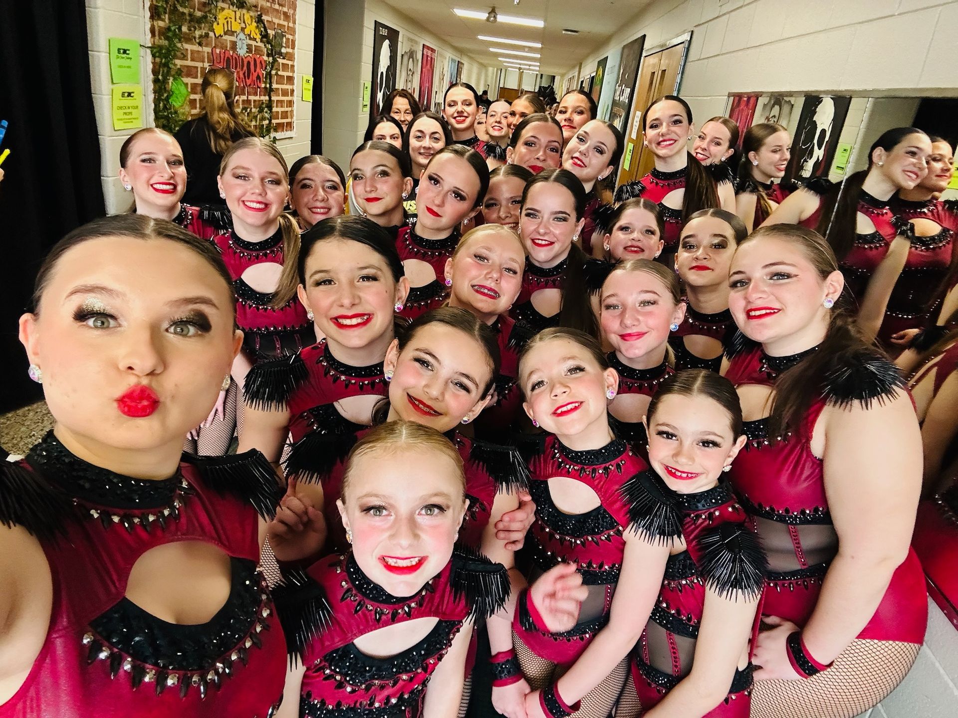 A group of young female dancers in matching red and black costumes pose for a selfie in a hallway. Most smile, a few pucker their lips.