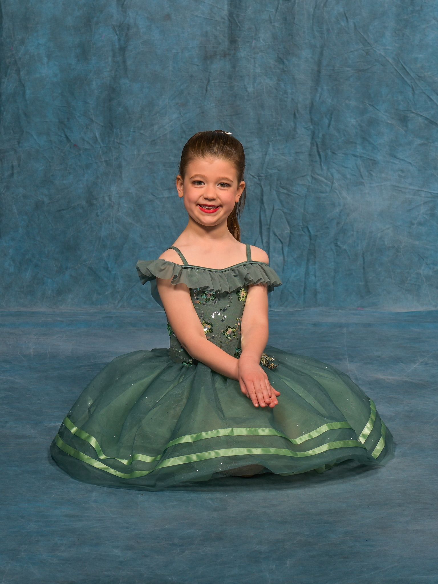 Young girl in a green dance dress with a tutu, smiling, seated on a blue backdrop.